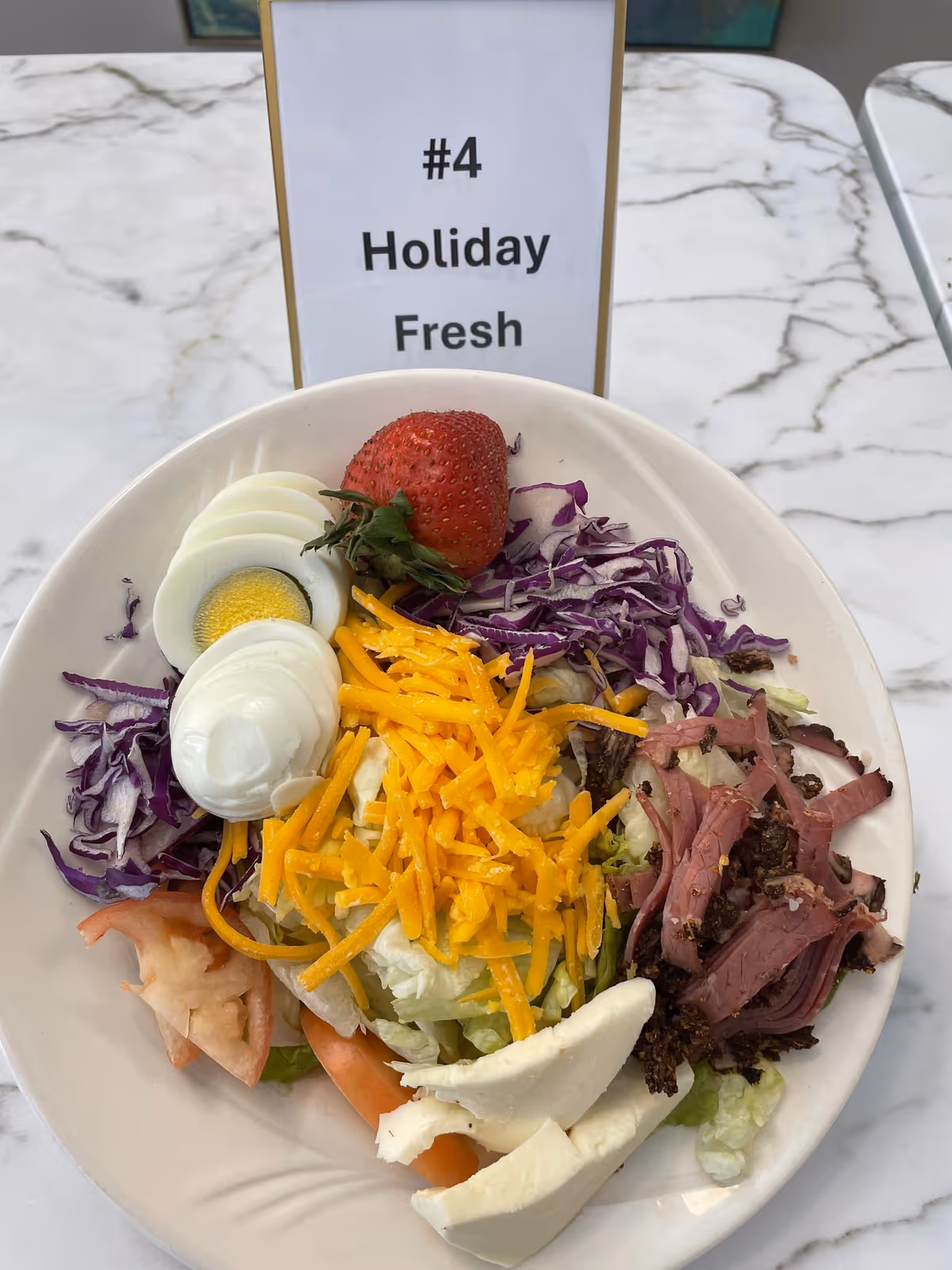 A plate of salad with shredded cheddar cheese, sliced hard-boiled egg, a strawberry, shredded purple cabbage, sliced tomato, slices of white cheese, and shredded meat on a bed of lettuce. Behind the plate is a sign that reads '#4 Holiday Fresh' on a marble-patterned table.
