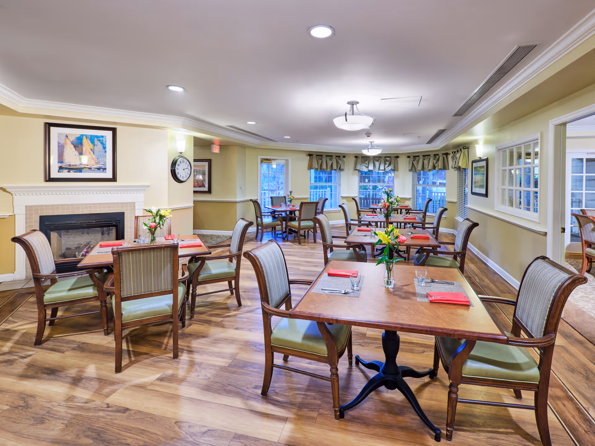 A bright and inviting dining room with wooden floors, several tables set with placemats, glasses, and red napkins. Each table has a small vase with fresh flowers. The room features a fireplace with a framed painting above it, a wall clock, and multiple windows with valances allowing natural light to enter.