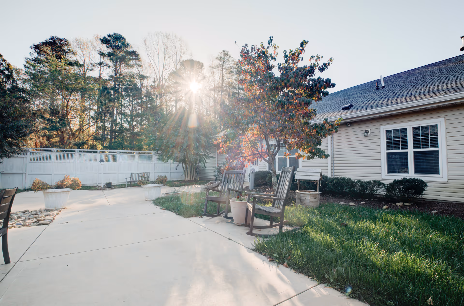 Outdoor patio area at Falls River Court Memory Care with wooden chairs, potted plants, a tree with autumn leaves, a white fence, and a building with beige siding and windows. Sunlight is shining through the trees in the background.