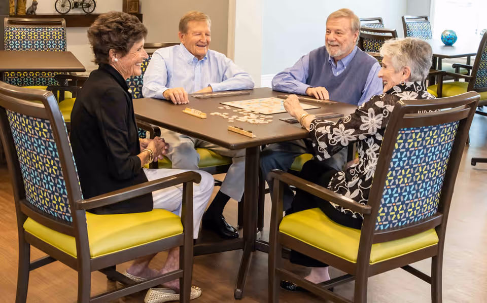 Four elderly people sitting around a square table playing a board game in a well-lit room with patterned chairs and wooden flooring.