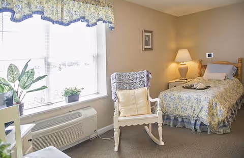 A cozy bedroom with a single bed covered in floral bedding, a white rocking chair with cushions, a bedside table with a lamp, a framed picture on the wall, and a window with floral valance letting in natural light. There is also a potted plant on the windowsill and an air conditioning unit below the window.