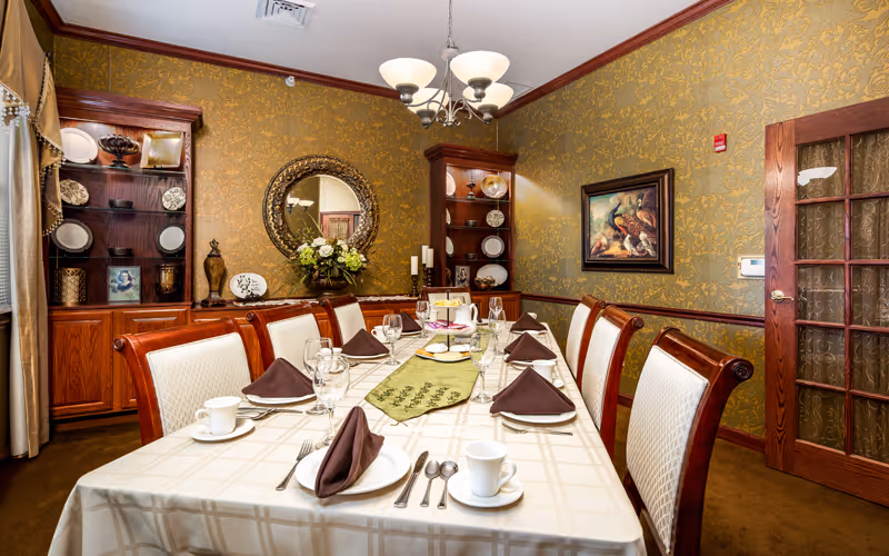 A formal dining room with a long table set for eight people. The table is covered with a beige checkered tablecloth and has a green runner with a floral design. Each place setting includes a white plate, a folded brown napkin, a cup and saucer, and silverware. The room features ornate gold wallpaper, wooden cabinets with decorative plates and vases, a round mirror with a floral arrangement beneath it, a chandelier overhead, and a framed painting of peacocks on the wall. There is a wooden door with glass panels on the right side.