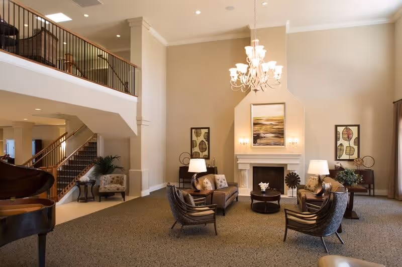 A spacious, elegant living room area with high ceilings and a large chandelier. The room features a fireplace with a painting above it, two sofas, two armchairs, side tables with lamps, and decorative plants. A grand piano is visible on the left side near a staircase with wooden railings leading to an upper floor.