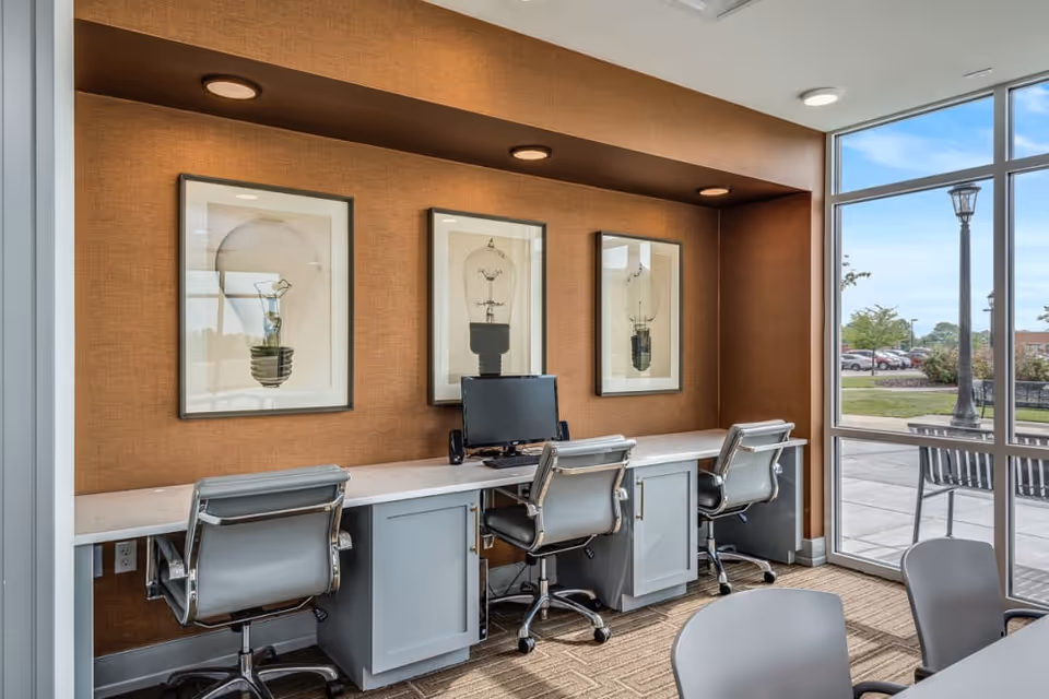 A modern workspace area with three gray office chairs in front of a long white desk with a computer monitor. The wall behind the desk is brown and decorated with three framed pictures of light bulbs. Large windows on the right side provide a view of an outdoor area with benches, a lamp post, and parked cars.