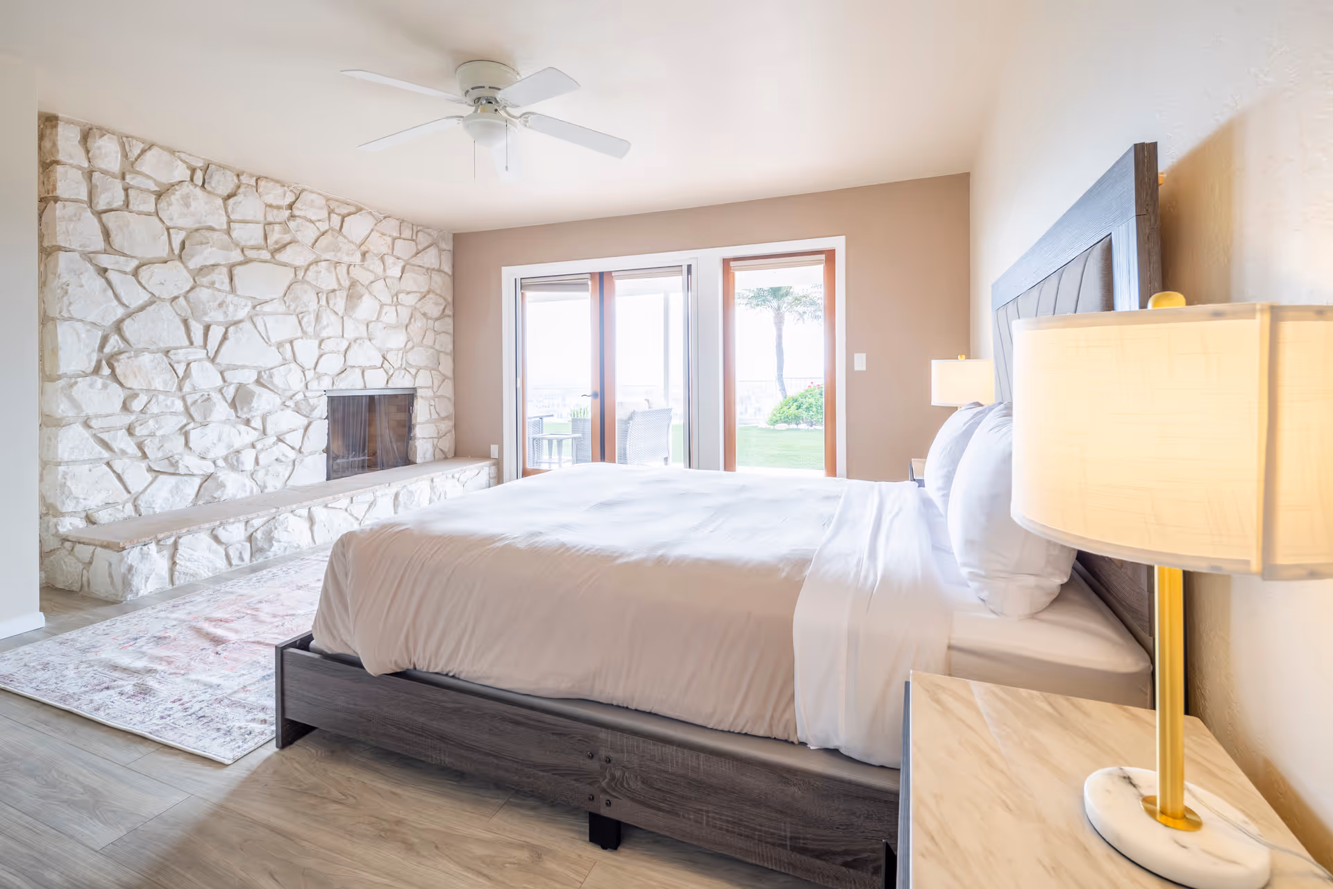 Bright bedroom with a large bed featuring white bedding, a wooden headboard, and a bedside table with a lamp. The room has a stone fireplace on one wall, a ceiling fan, and sliding glass doors leading to an outdoor area with greenery.