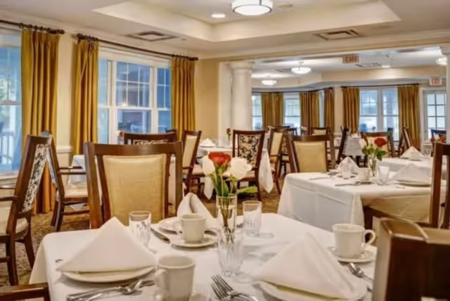 A well-lit dining room with multiple tables covered in white tablecloths, each set with white napkins, cups, glasses, and silverware. Chairs with wooden frames and cushioned seats surround the tables. Yellow curtains frame the windows, and there are white columns and ceiling lights in the room.
