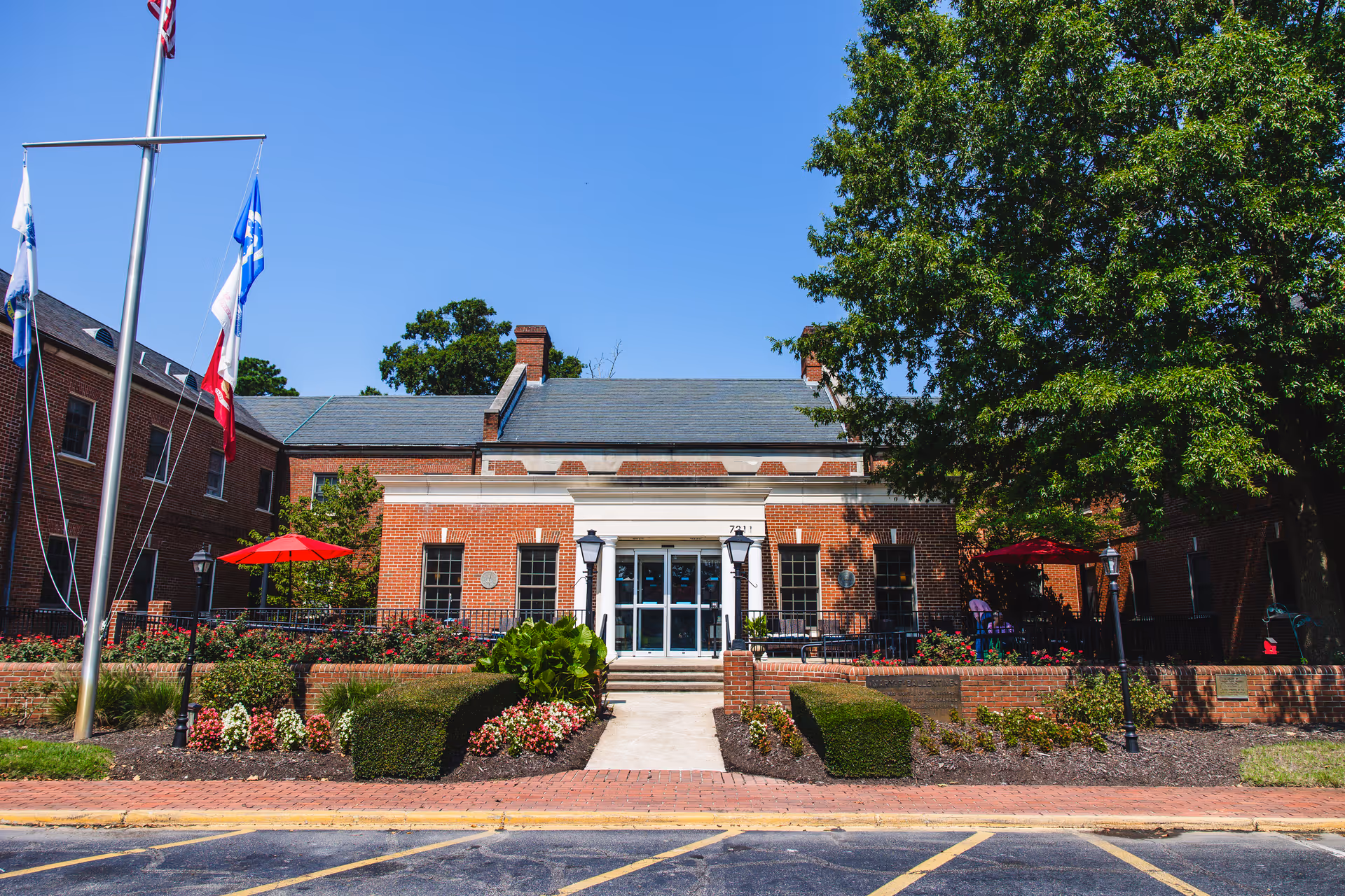Front exterior view of a brick building with a slate roof, surrounded by well-maintained landscaping including bushes, flowers, and trees. There are flagpoles with flags on the left side and red umbrellas providing shade over outdoor seating areas. The entrance has glass double doors with two lamp posts on either side.