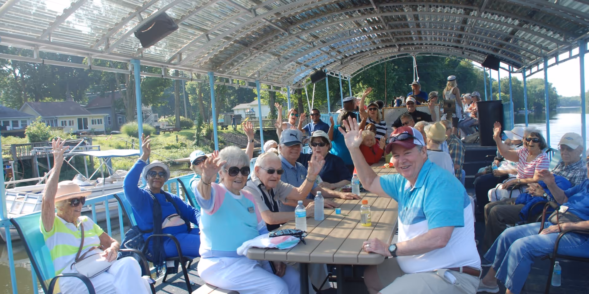 A group of elderly people sitting around a long table on a covered boat deck, smiling and waving at the camera. The boat is on a river or lake with trees and houses visible in the background. The weather is sunny and bright.