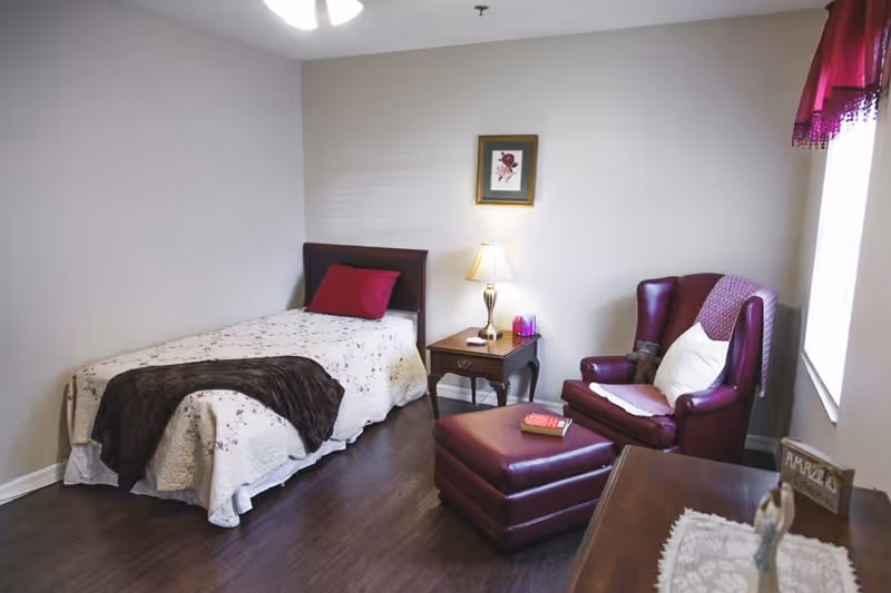 A cozy senior living bedroom with a single bed covered in a floral quilt and a dark throw blanket. Next to the bed is a wooden nightstand with a lamp and some small items. A burgundy armchair with a white pillow and a matching ottoman are placed near a window with a red valance. The room has wooden flooring and neutral-colored walls with a framed floral picture hanging above the bed.