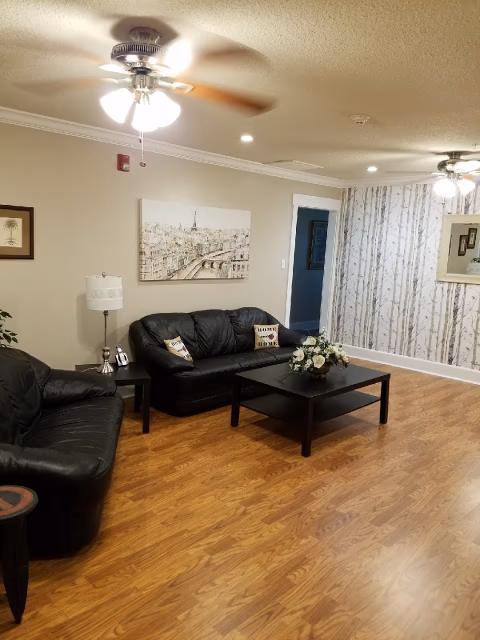 Cozy living room with black leather sofas around a coffee table topped with a floral arrangement, wood-look floors, wall art, and ceiling fans.
