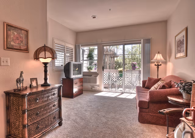 A cozy living room with a red sofa, a wooden side table with a lamp, a wooden chest of drawers with a decorative lamp and small horse figurine, a TV on a wooden stand, and large sliding glass doors leading to a balcony with outdoor plants.