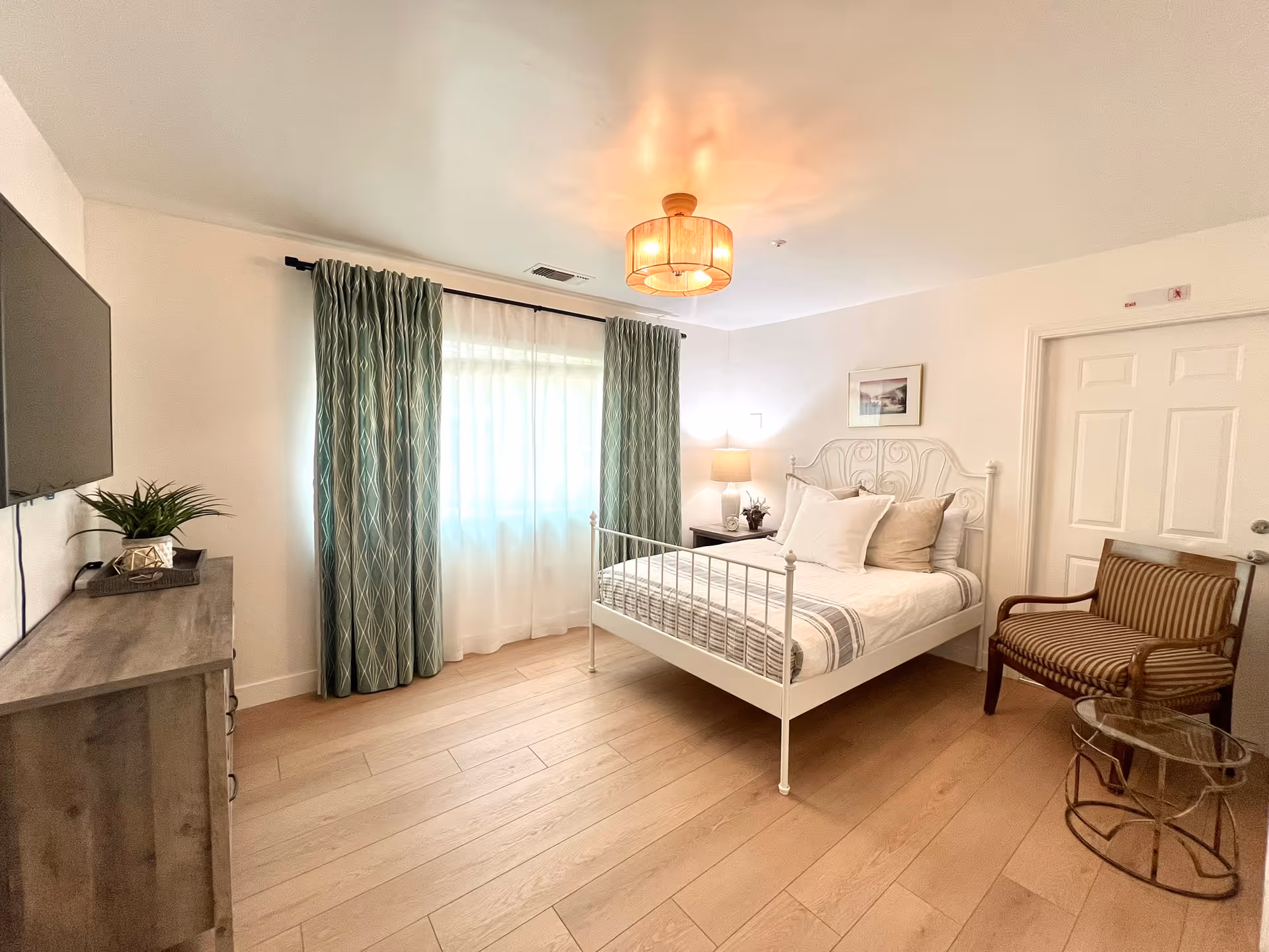 Sunlit bedroom with a white metal bed, striped bedding, dresser, armchair, and green curtains.