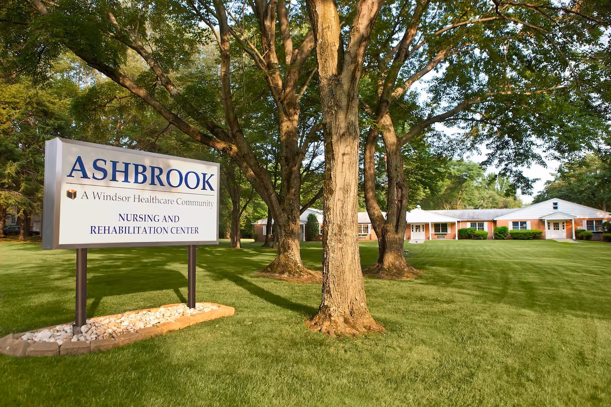 Outdoor view of Ashbrook Nursing and Rehabilitation Center with a large sign in the foreground and a single-story brick building surrounded by green grass and trees in the background.