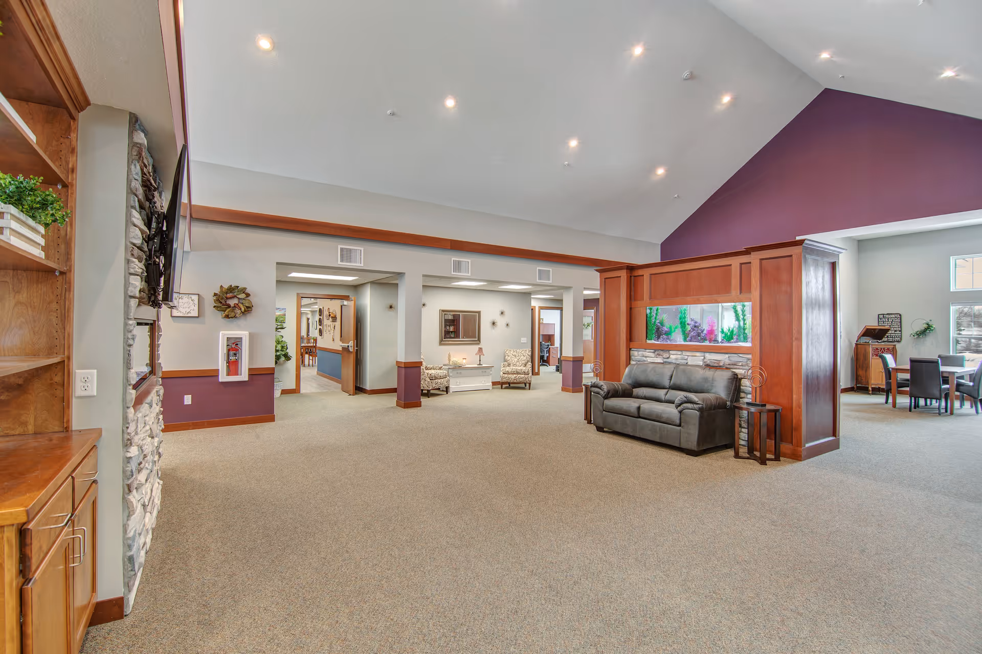 Spacious common lounge in a senior living facility with a sofa in front of a built-in aquarium, high vaulted ceiling, and adjacent dining area.