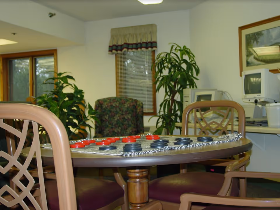 Round table with a checkers game and chairs in a senior community common room with plants and computers in the background.