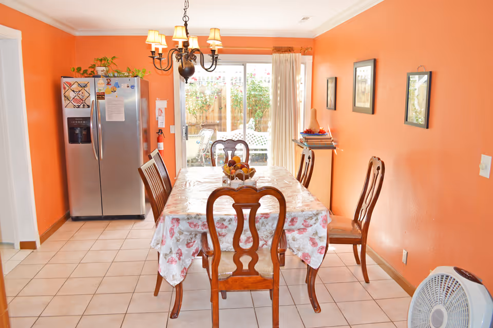 A dining room with orange walls and tiled floor featuring a wooden dining table covered with a floral tablecloth and six wooden chairs. A chandelier hangs above the table. There is a stainless steel refrigerator on the left side, a small cabinet with books and decorative items on the right, and a sliding glass door leading to an outdoor patio with plants visible outside.