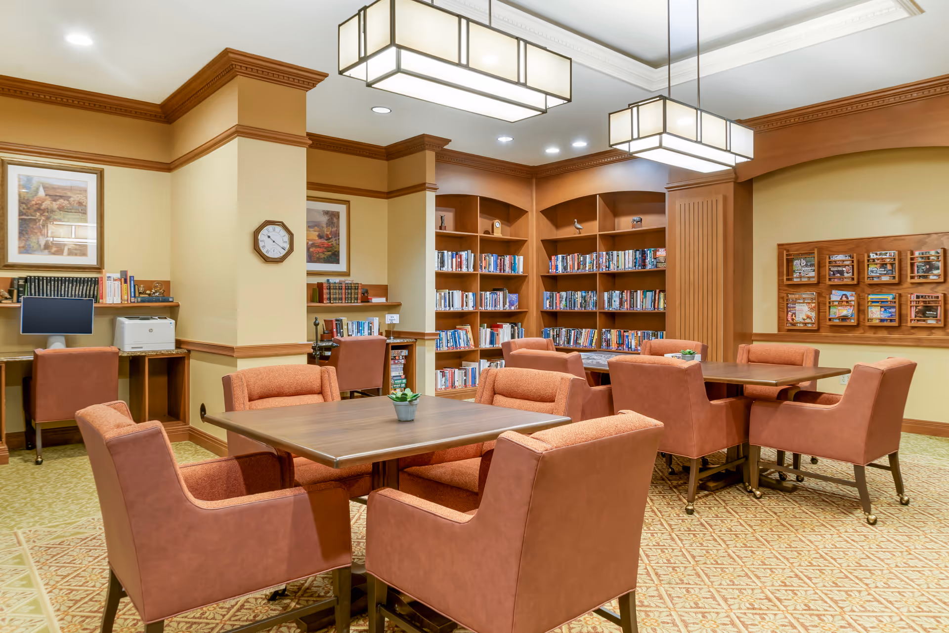 A cozy library and seating area in a senior living facility with wooden bookshelves filled with books, two tables surrounded by cushioned chairs, a computer workstation with a printer, framed artwork on the walls, and warm lighting from rectangular ceiling fixtures.