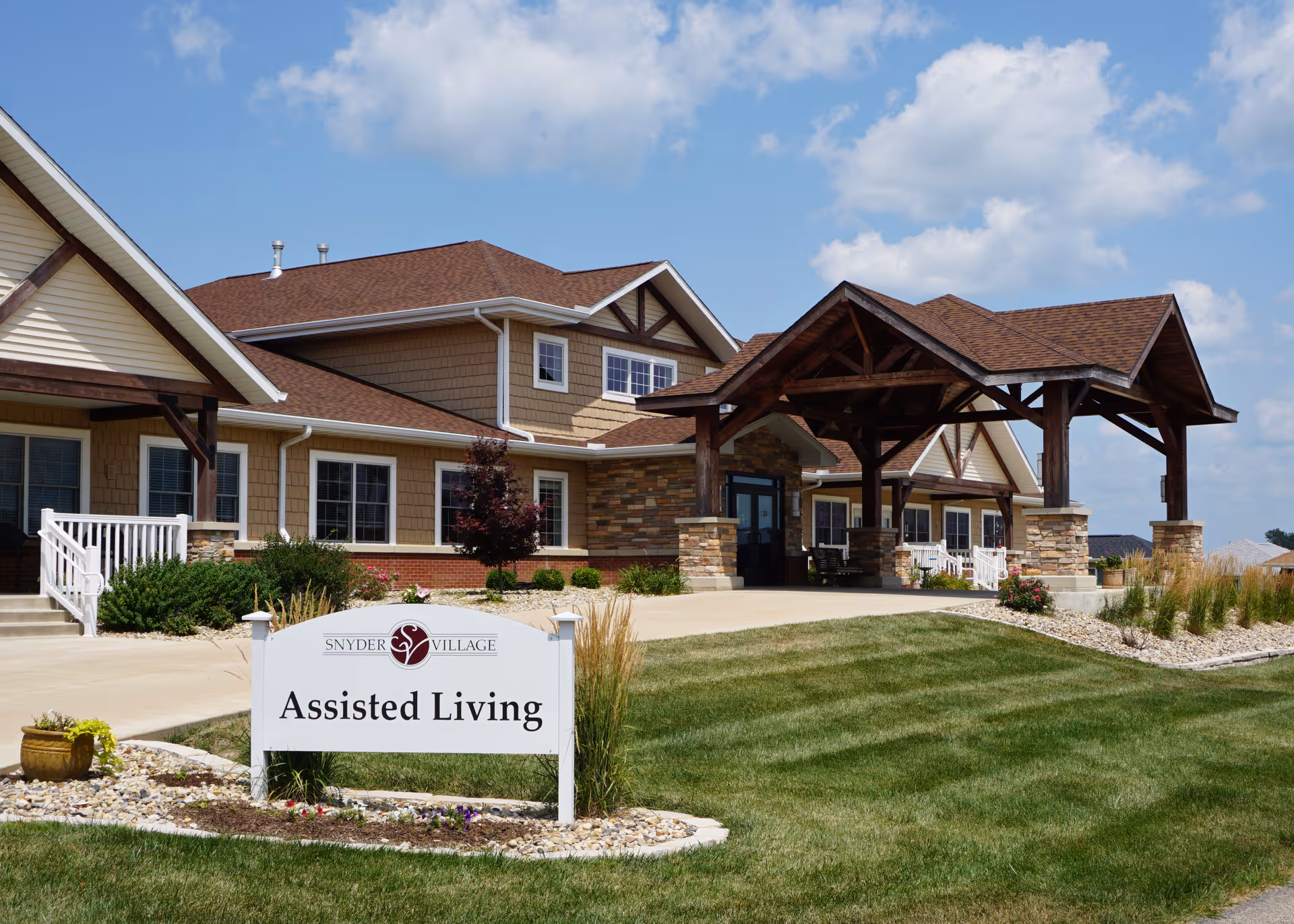 Exterior view of Snyder Village Assisted Living facility showing a large building with a covered entrance, stone and wood accents, and a well-maintained lawn under a partly cloudy sky.
