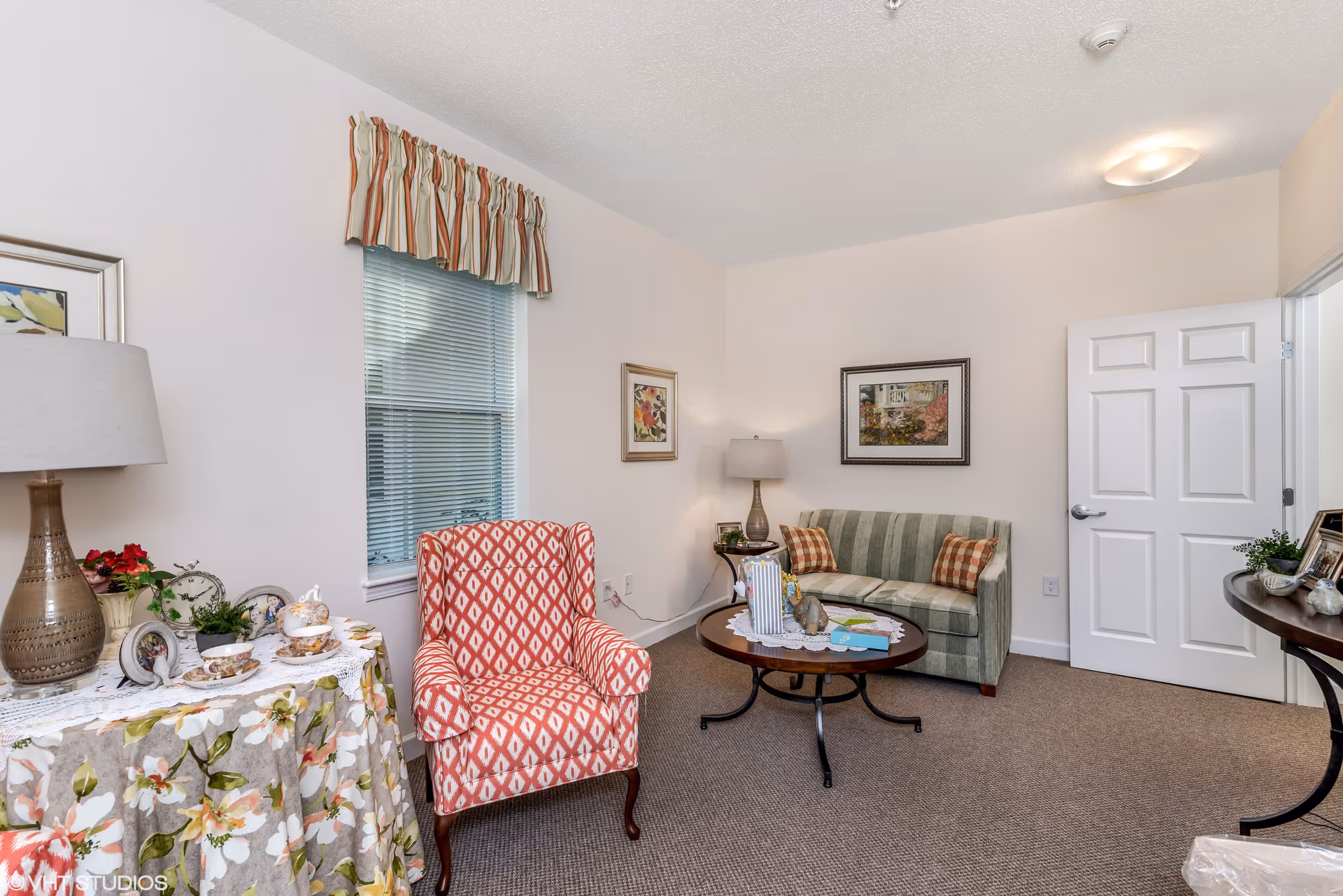 A cozy living room with a striped green loveseat, a red patterned armchair, a round wooden coffee table with decorative items, a side table with a lamp and floral tablecloth, framed artwork on the walls, and a window with blinds and a striped valance.