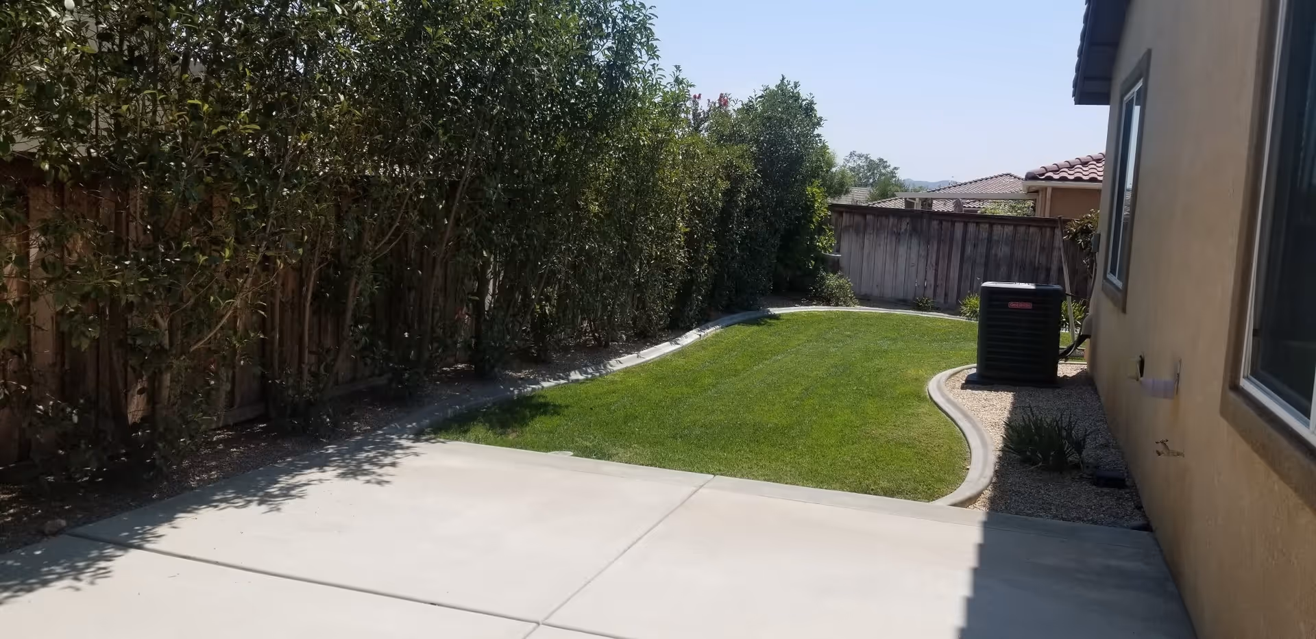 A backyard area with a concrete patio in the foreground, a small curved lawn bordered by a concrete edge, tall green bushes along a wooden fence on the left, and the side of a beige house with windows and an air conditioning unit on the right under a clear blue sky.