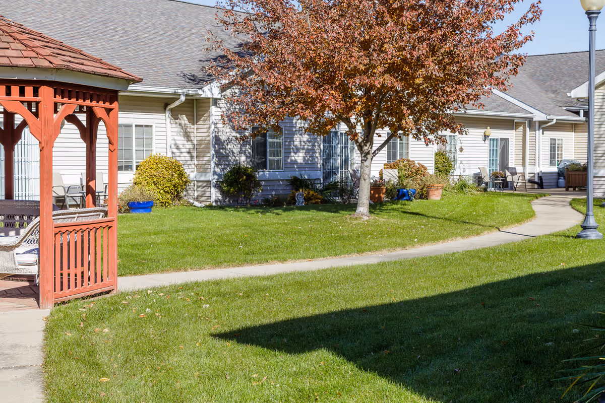 Outdoor view of a senior living facility courtyard with a red wooden gazebo on the left, a tree with autumn-colored leaves in the center, green grass, a curved concrete pathway, and beige buildings with windows and doors in the background.