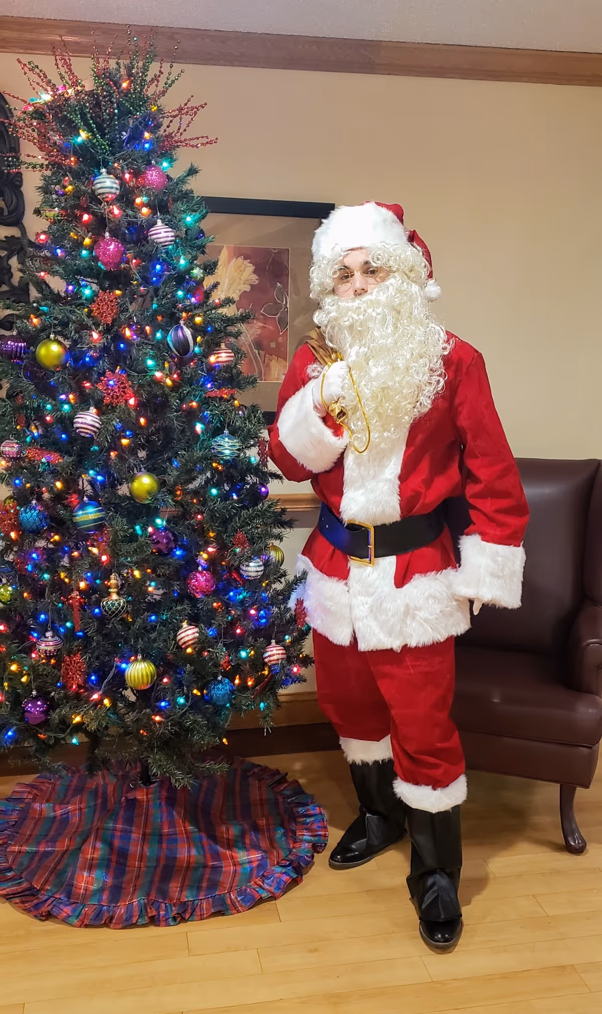 A person dressed as Santa Claus stands next to a decorated Christmas tree with colorful lights and ornaments in a room with wooden flooring and a brown armchair.