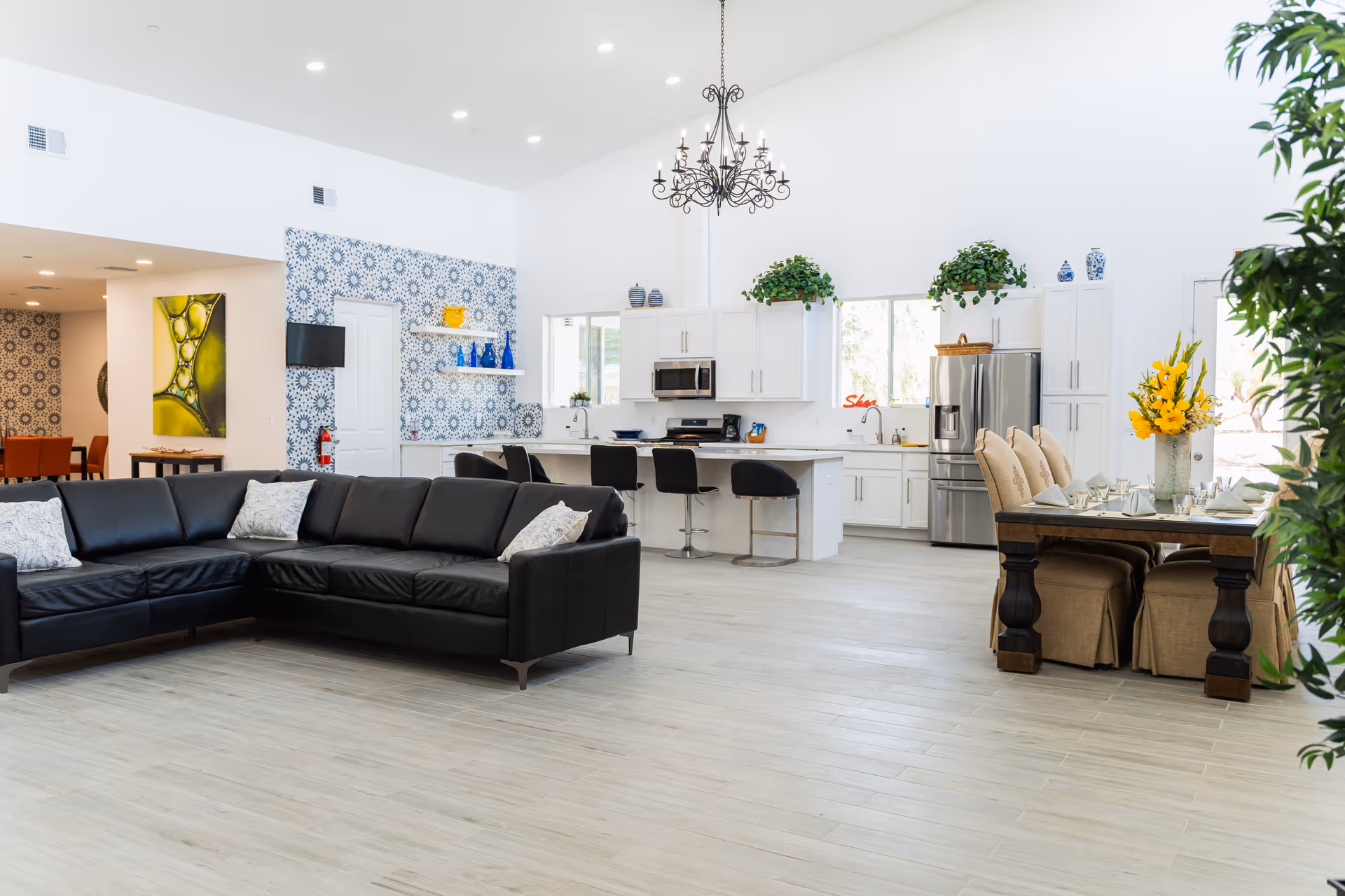 Bright and spacious open-concept living area featuring a black leather sectional sofa, a modern kitchen with white cabinetry, stainless steel appliances, and a breakfast bar with black stools. A dining table with beige upholstered chairs and a vase of yellow flowers is set near large windows. The room has light wood flooring, white walls, decorative blue patterned wallpaper on one wall, and a wrought iron chandelier hanging from the ceiling.