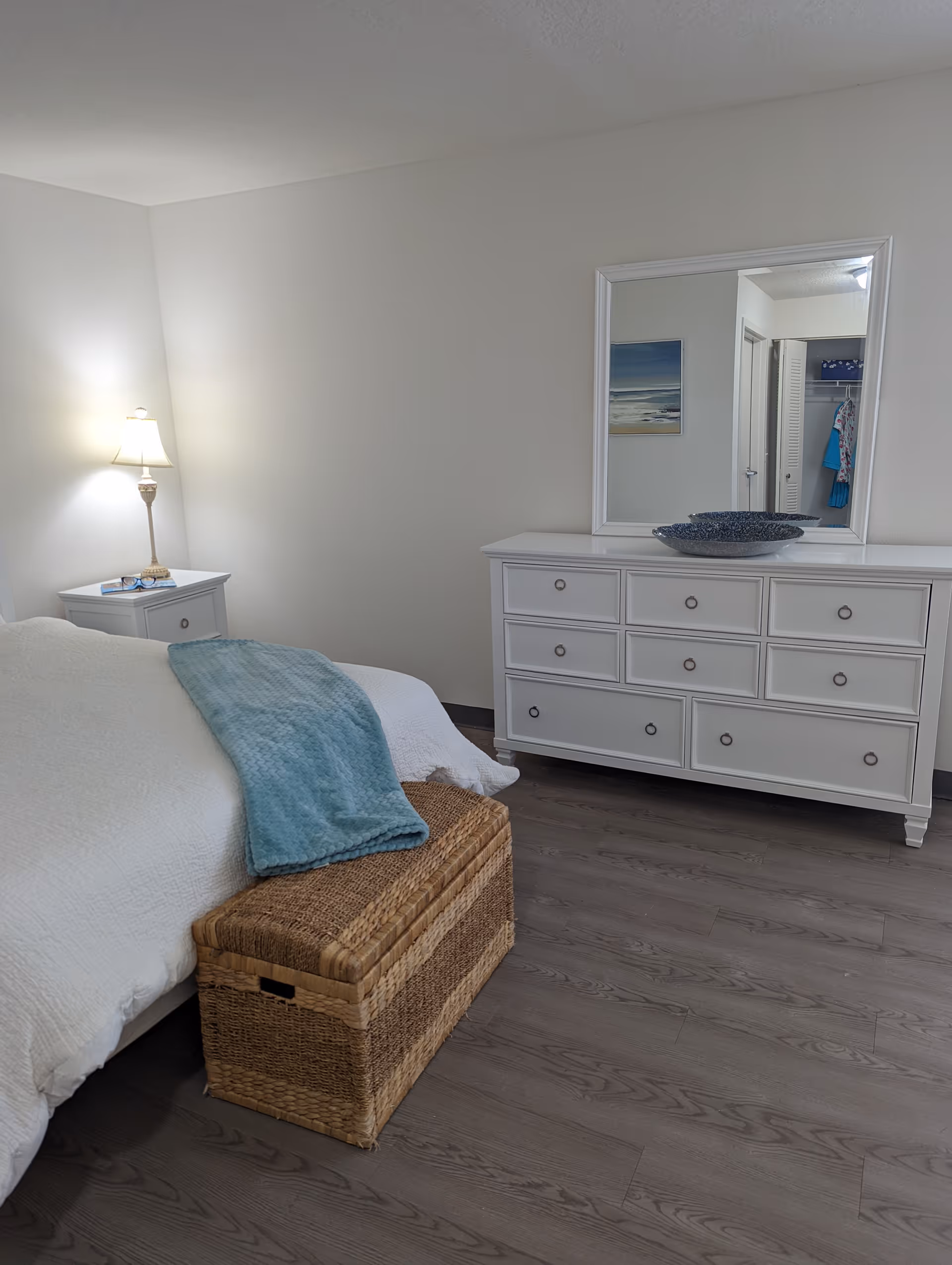 A bedroom with a white bed covered with a white quilt and a light blue throw blanket. There is a wicker storage chest at the foot of the bed. To the left, a white nightstand with a lamp and some items on it. On the right side, a white dresser with multiple drawers and a large mirror above it. The floor is a light wood laminate, and the walls are painted white.