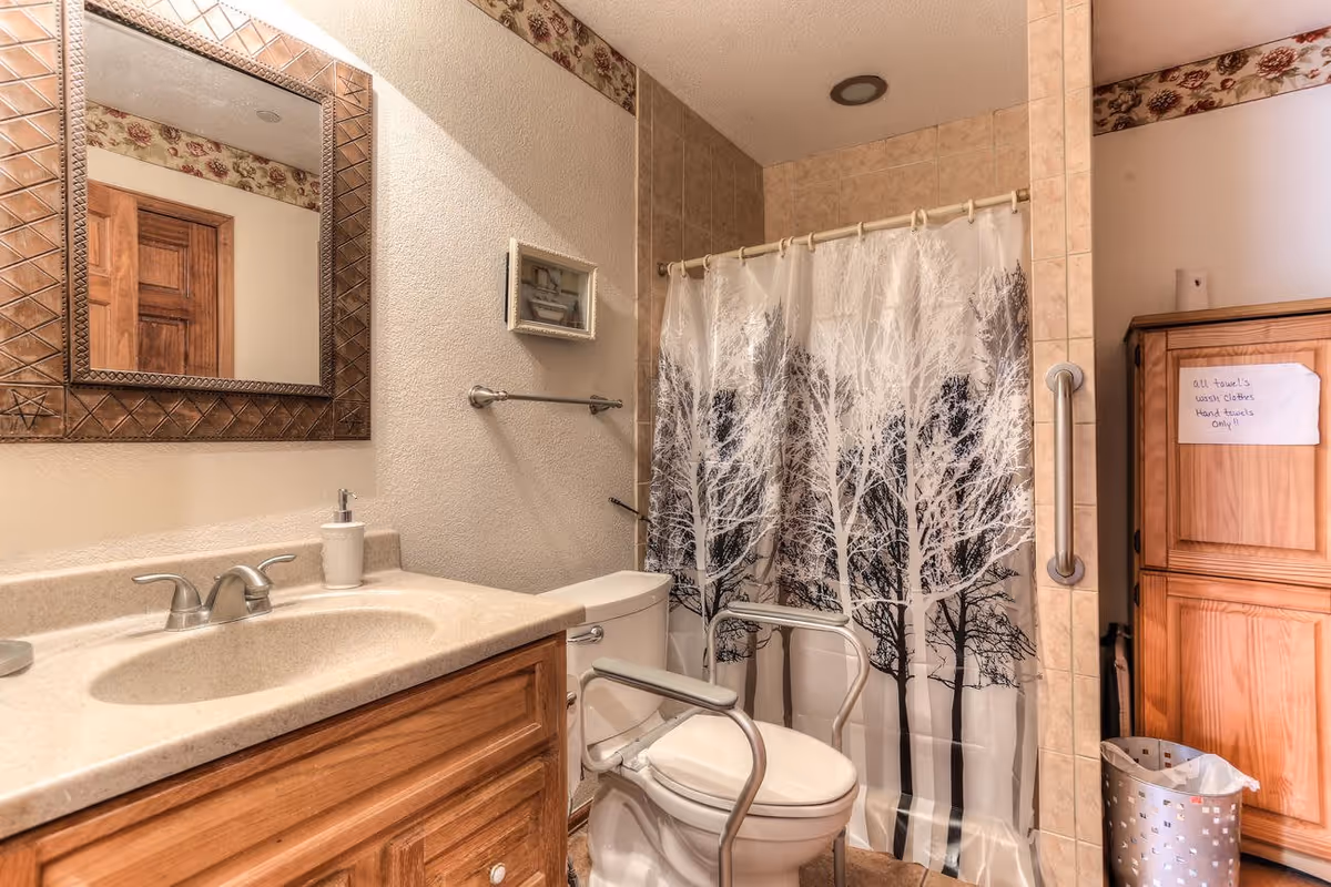 Bathroom with a wooden vanity and beige countertop, a large decorative mirror above the sink, a toilet with safety rails, a shower with a curtain featuring a tree design, and a wooden cabinet with a handwritten note on it.