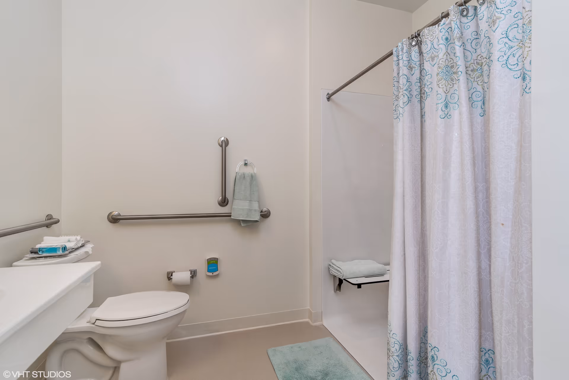 A clean and accessible bathroom featuring a toilet with grab bars on the walls, a towel hanging on a ring, a shower area with a patterned curtain and a fold-down seat, and a light blue bath mat on the floor.