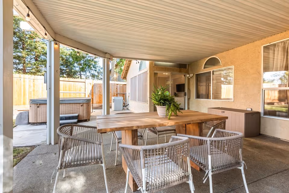 Covered outdoor patio area with a wooden table and six woven chairs arranged around it. A potted plant is placed on the table. In the background, there is a hot tub, a wooden fence, and windows of the building.