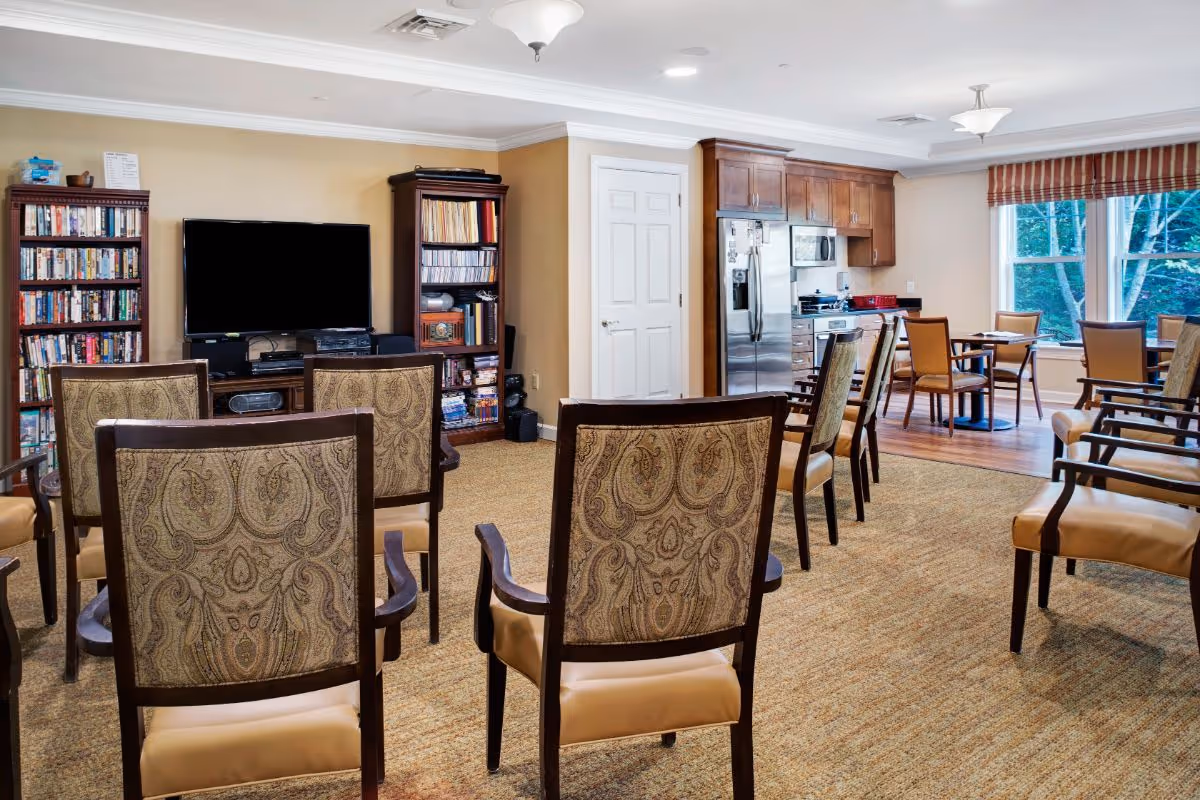 Common living area in a senior facility with rows of upholstered chairs facing a television, bookshelves, and a kitchenette with dining tables by a window.