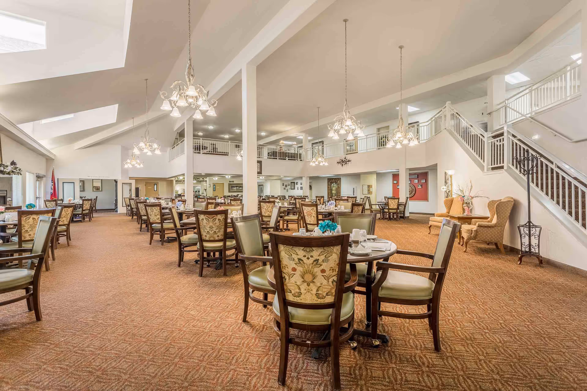Spacious dining room in a senior living facility with multiple round tables and chairs arranged neatly. The room features high ceilings with skylights, elegant chandeliers, carpeted floors, and a staircase leading to an upper level. There are decorative armchairs and a coat rack near the staircase.