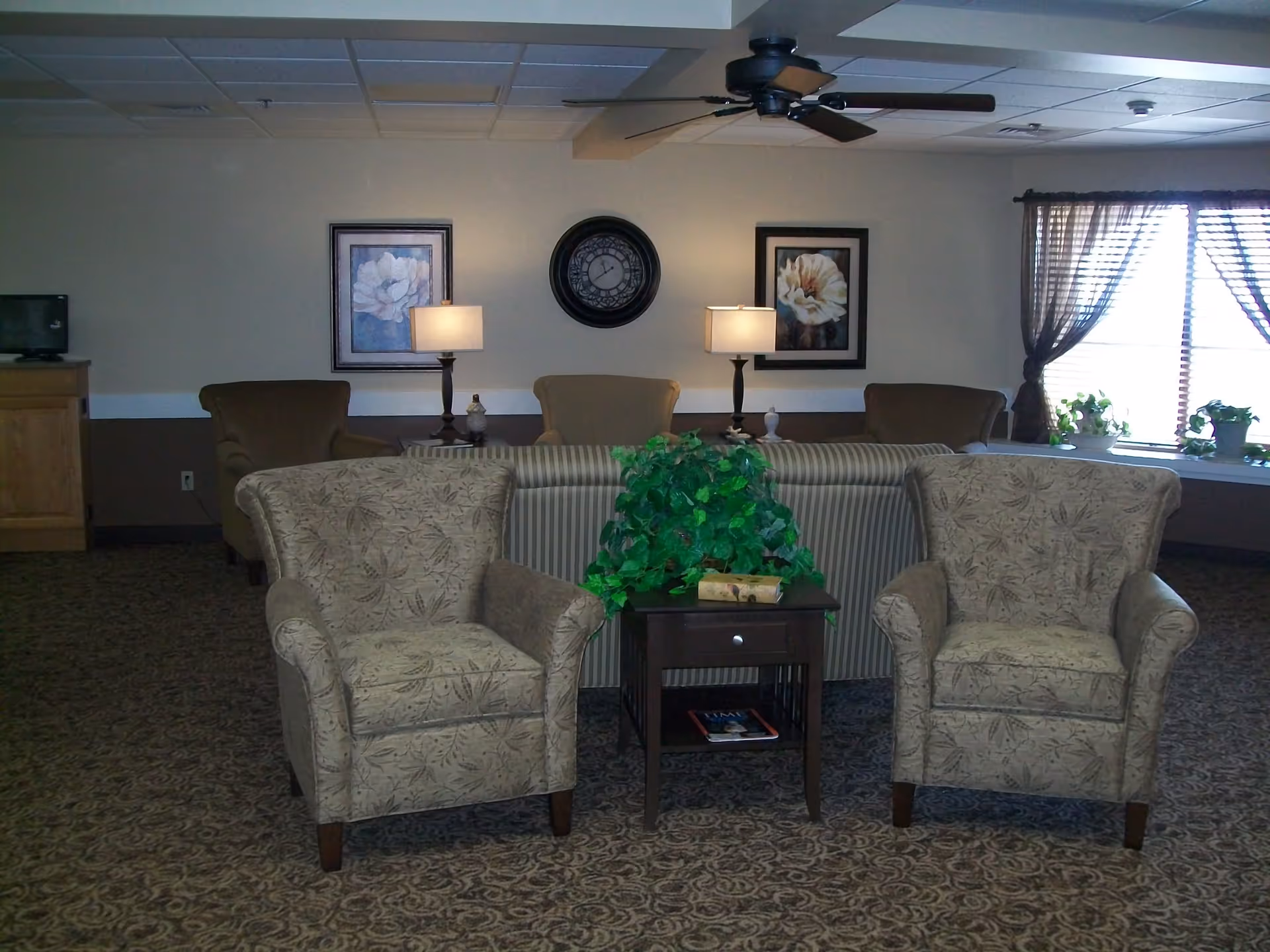 Comfortable common room with two patterned armchairs, a small side table with a plant, a striped sofa, lamps, wall art, and a window with curtains.