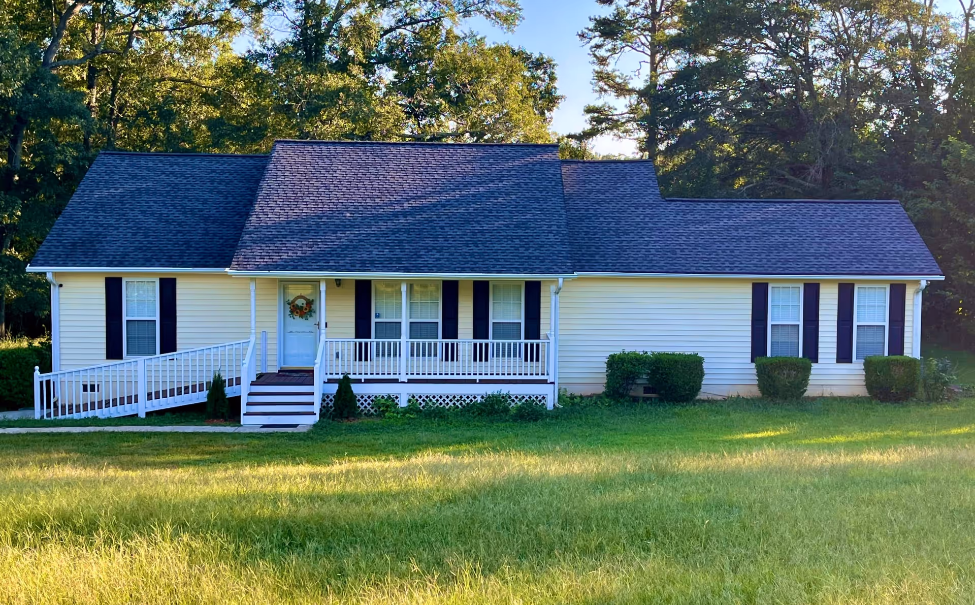 Single-story light yellow house with a dark shingled roof, white porch railing, and a wheelchair ramp leading to the front door. The house is surrounded by green grass and trees in the background.