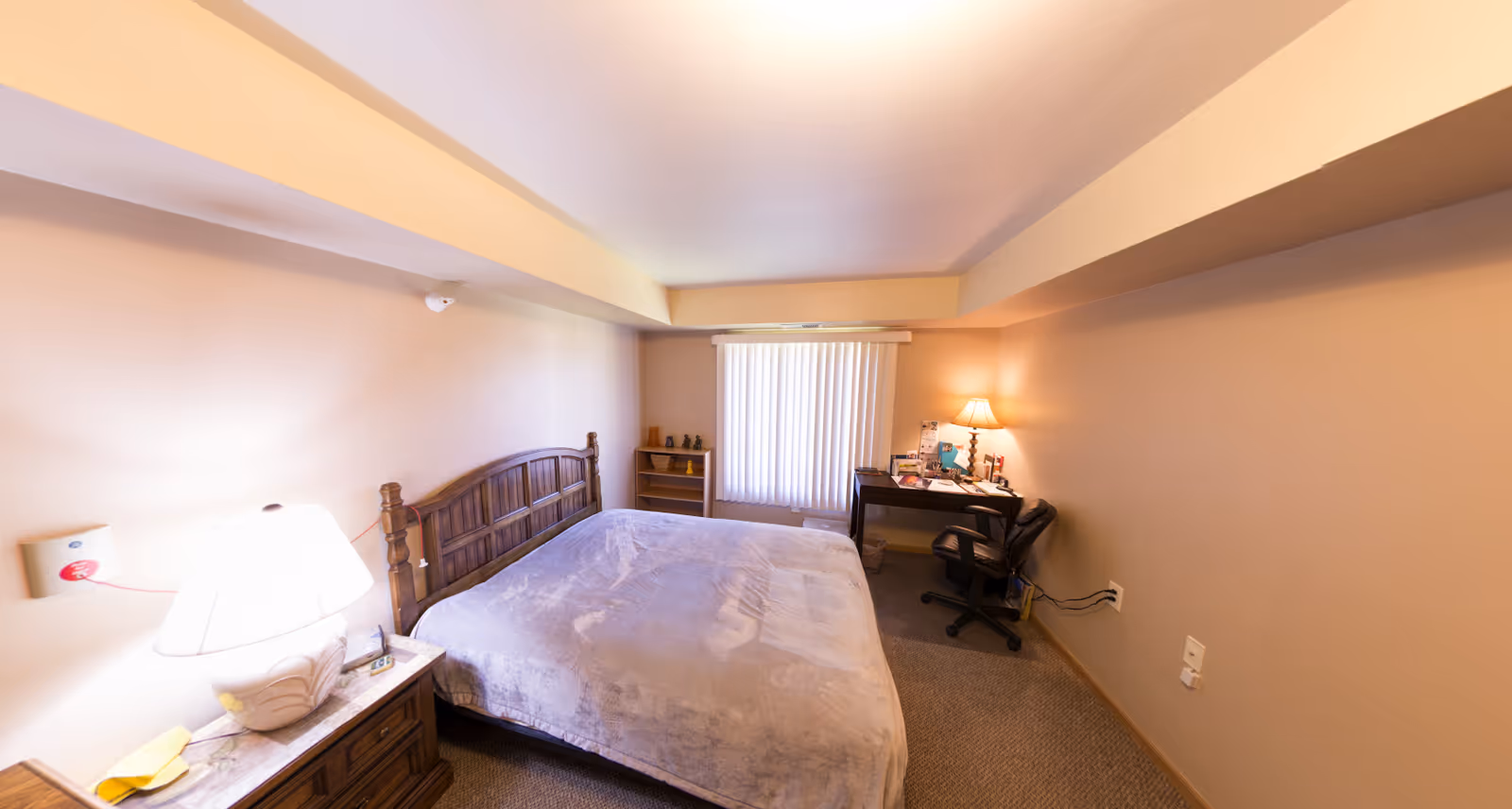 A bedroom in a retirement community featuring a large bed with a wooden headboard, a bedside table with a lamp, a small bookshelf, a desk with a lamp and various items, and a black office chair. The room has beige walls, carpeted floor, and a window with vertical blinds.