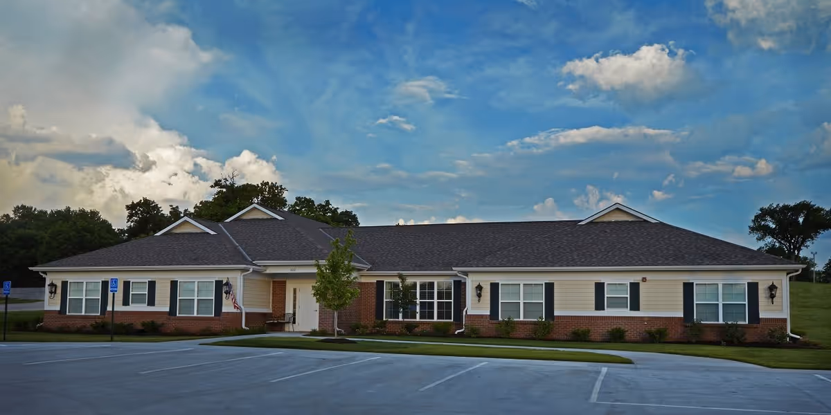 Single-story building with beige siding and red brick accents, multiple windows with black shutters, a dark shingled roof, and a small front porch with an American flag. The building is surrounded by a well-maintained lawn and a parking lot with marked spaces, including handicapped spots. The sky above is partly cloudy with blue patches.