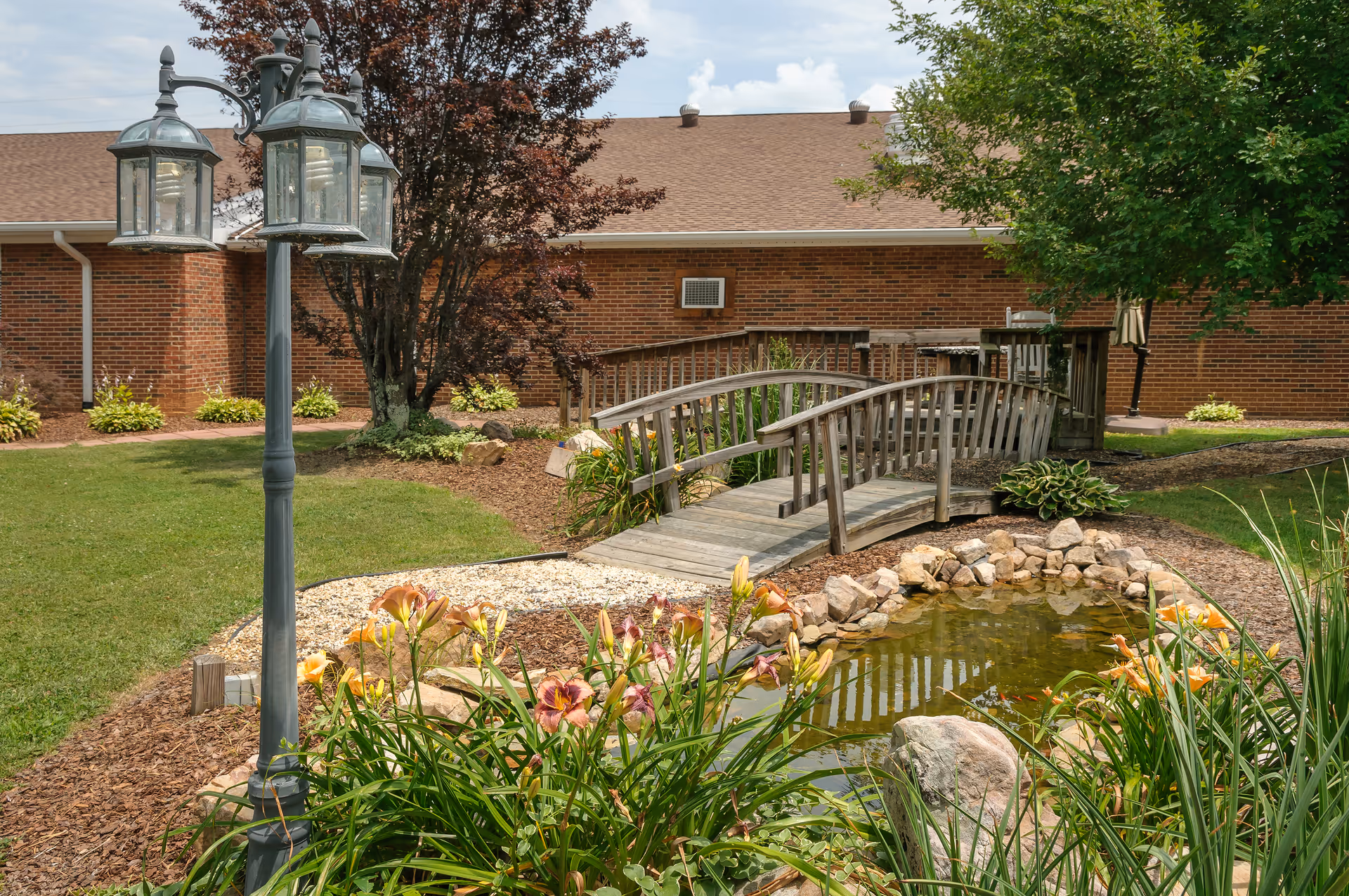 A landscaped courtyard with a small pond, wooden footbridge, flowering plants, and a lamp post in front of a brick building.