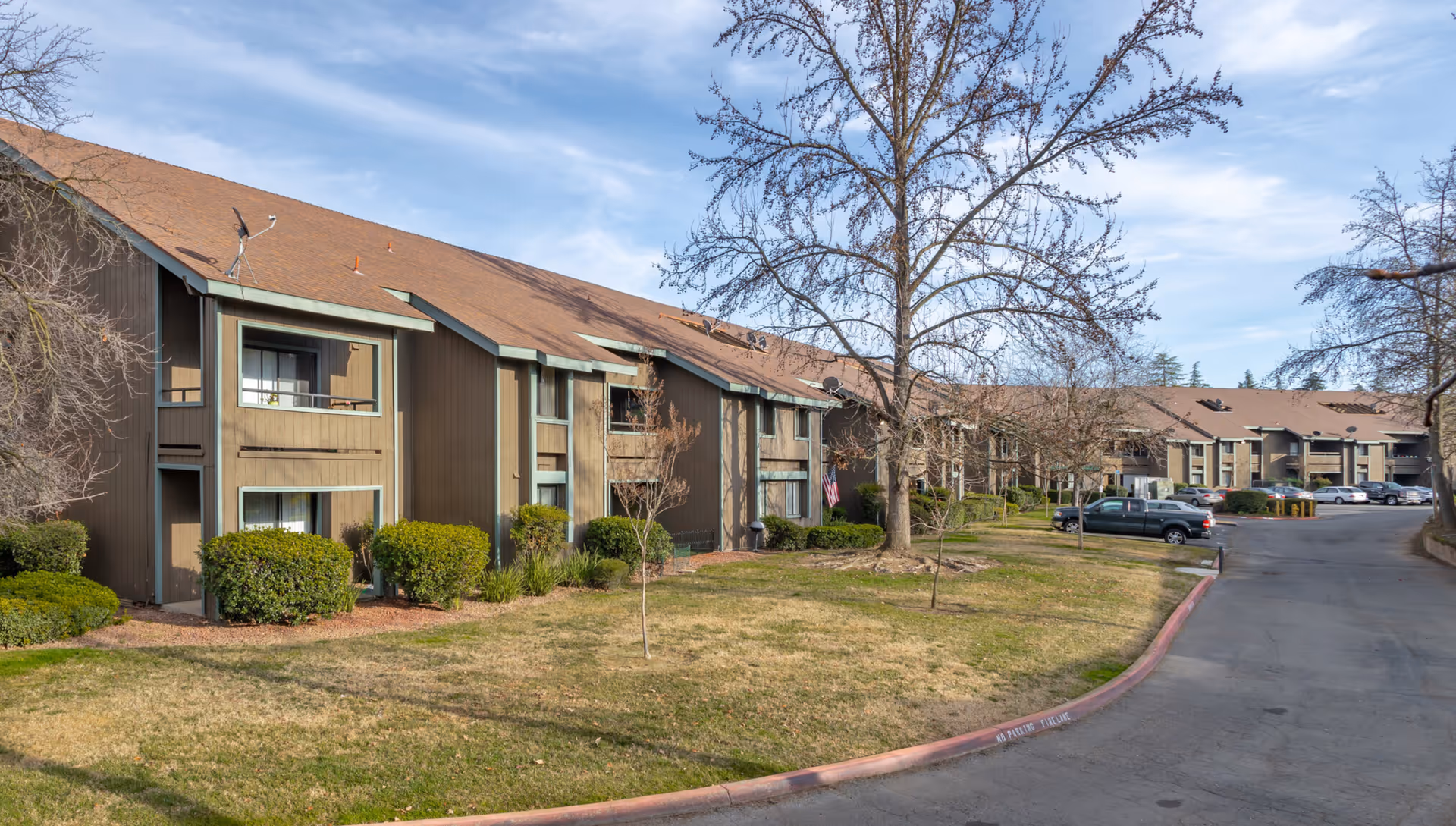 Exterior view of a senior living facility with brown buildings featuring green trim, a sloped roof, and multiple windows. There are leafless trees and trimmed bushes in front of the buildings, a grassy area, and a paved driveway with parked cars under a blue sky with some clouds.
