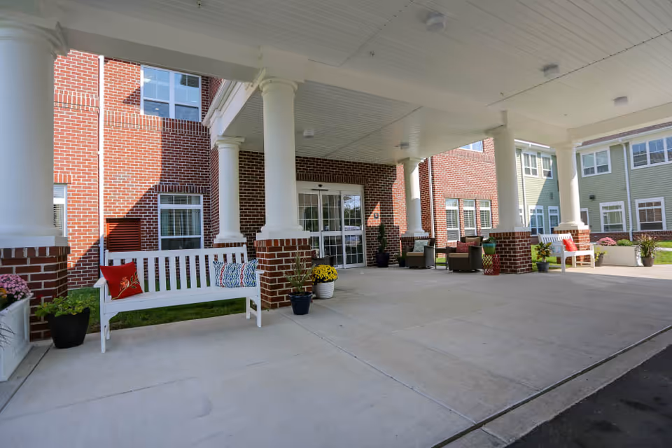 Covered entrance area of a senior living facility with white columns and brick walls. There are white benches with colorful cushions, potted plants, and outdoor seating near the glass entrance doors.