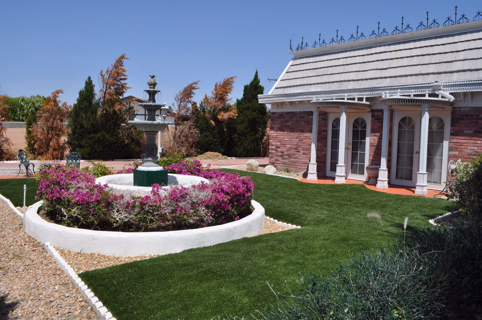 A courtyard with a central multi-tier fountain surrounded by purple flowers, manicured lawn, and a brick building with white columns.