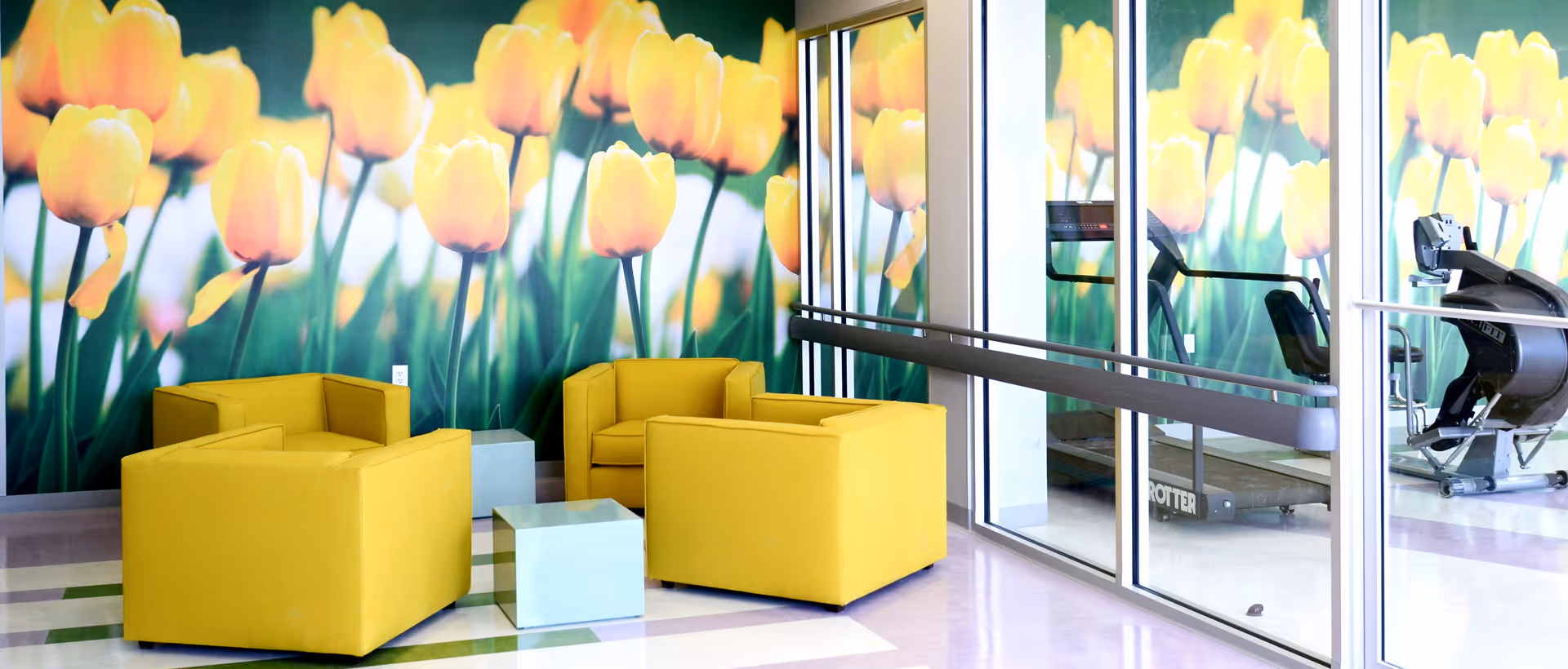 A bright and modern seating area with four yellow armchairs arranged around two small square tables. The background features a large wall mural of yellow tulips. To the right, there is a glass wall revealing exercise equipment including a treadmill and a stationary bike.