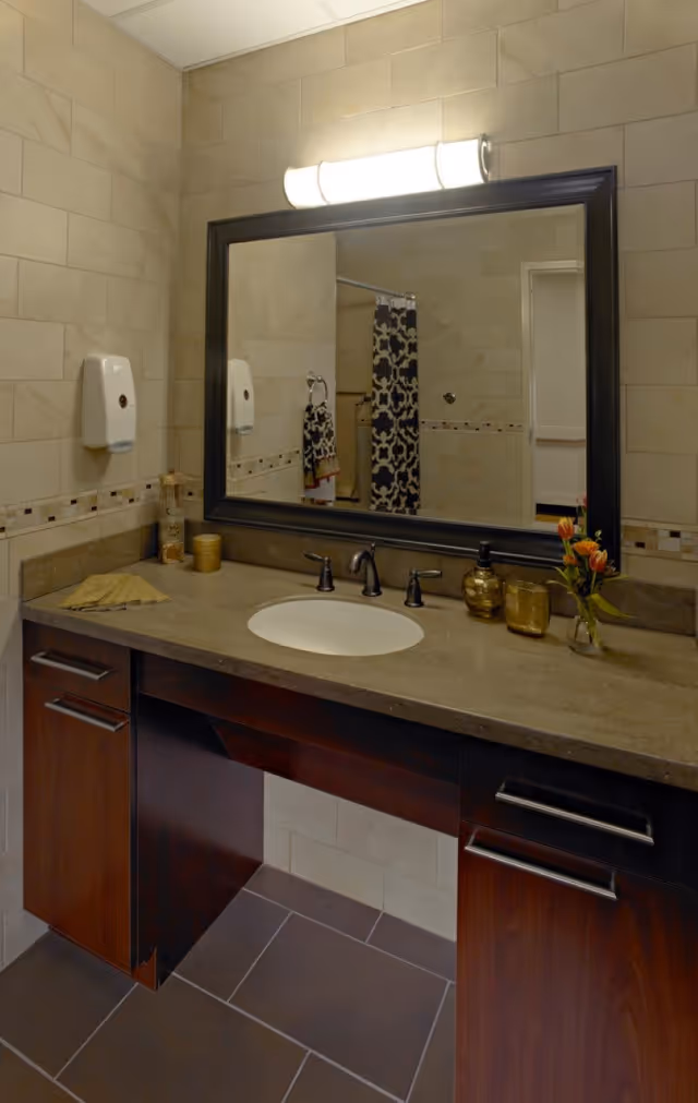 A bathroom vanity area with a large rectangular mirror framed in dark wood. The countertop is beige with a built-in sink and dark bronze faucet fixtures. On the countertop, there are decorative items including a small vase with orange flowers, a soap dispenser, and a candle holder. The walls are tiled in a light beige color with a decorative tile border. A towel ring with a patterned towel and a soap dispenser are mounted on the wall. The floor is tiled in dark brown.