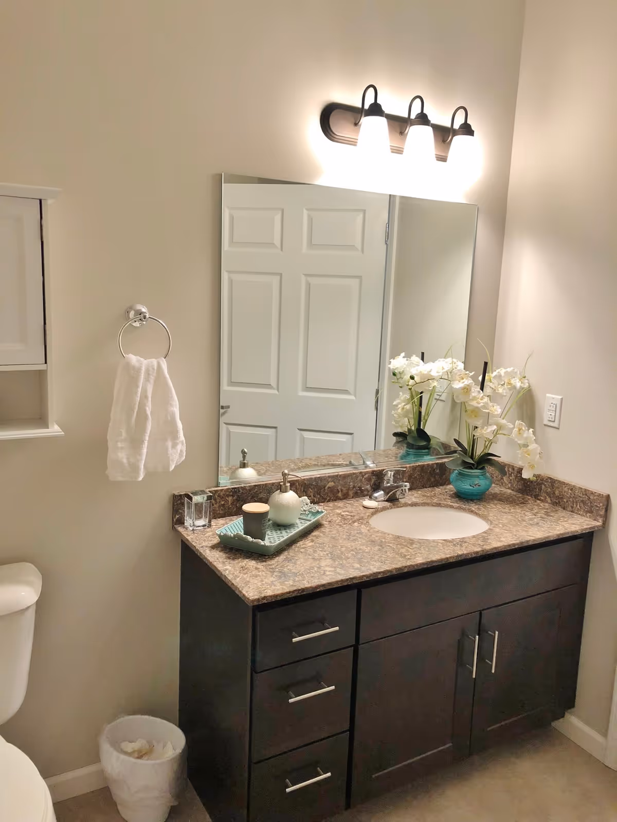 A clean and well-lit bathroom vanity area with a large mirror above a dark wood cabinet with a marble countertop. On the countertop, there is a green tray holding a soap dispenser and a small container, and a turquoise vase with white flowers. A white towel hangs on a ring on the wall next to the mirror, and a white door is reflected in the mirror. A toilet and a small trash bin are partially visible to the left.