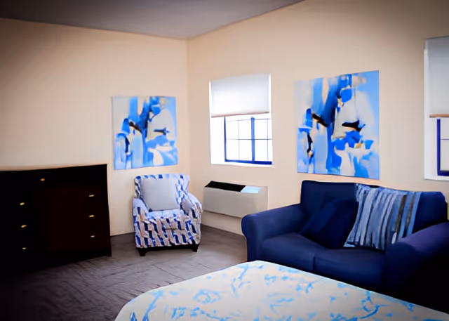 A cozy living room area with a blue patterned armchair, a dark blue sofa with striped cushions, two abstract blue and white paintings on the walls, a window with a white blind, a dark wooden dresser, and a bed with a blue and white patterned bedspread in the foreground.