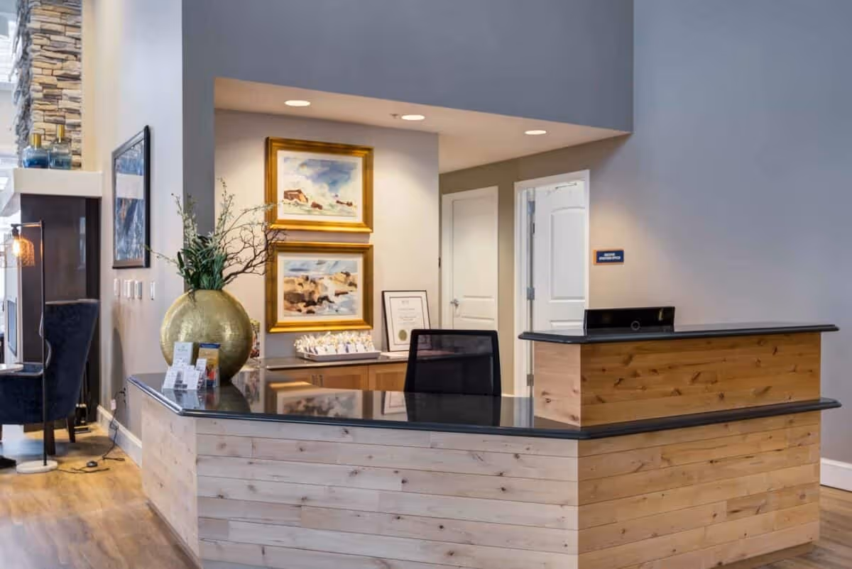 Reception desk area with a wood-paneled counter and glass top, decorative gold vase, framed paintings, and seating in a lobby.