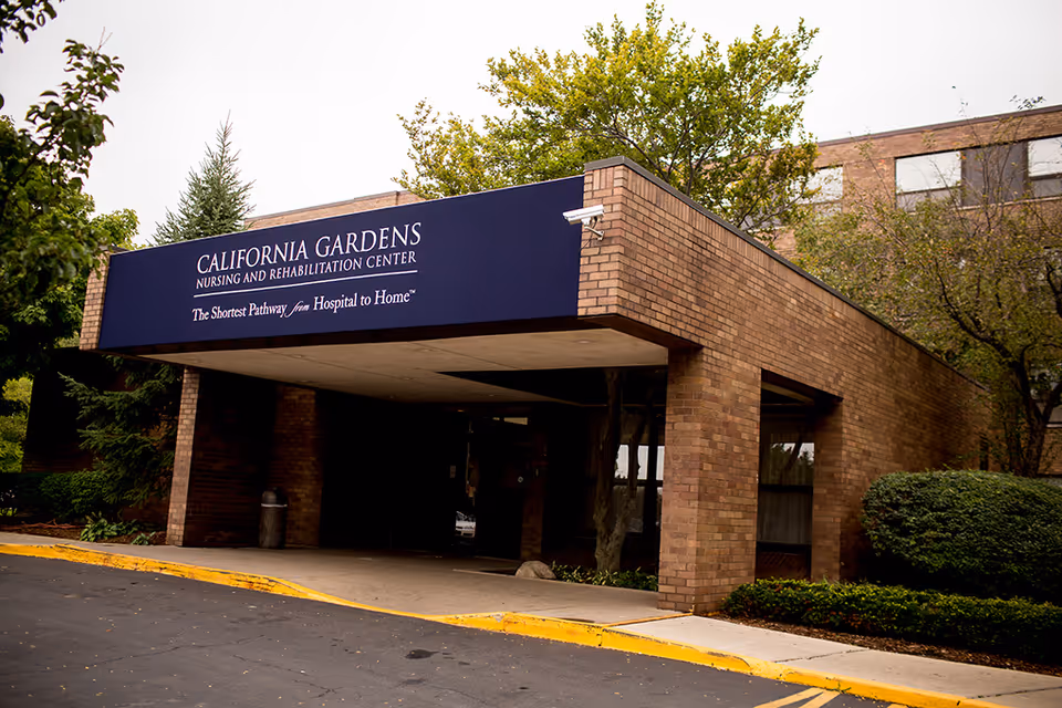 Entrance to California Gardens Nursing and Rehabilitation Center with a covered drop-off area, surrounded by trees and bushes, and a brick exterior building.