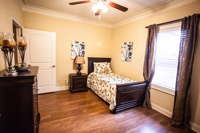 A neatly arranged bedroom with a single dark wood bed featuring a floral bedspread and matching pillow. There is a dark wood nightstand with a lamp beside the bed, and a matching dresser with decorative items on top. The room has light beige walls, wooden flooring, a ceiling fan with lights, and a window with dark curtains and blinds.