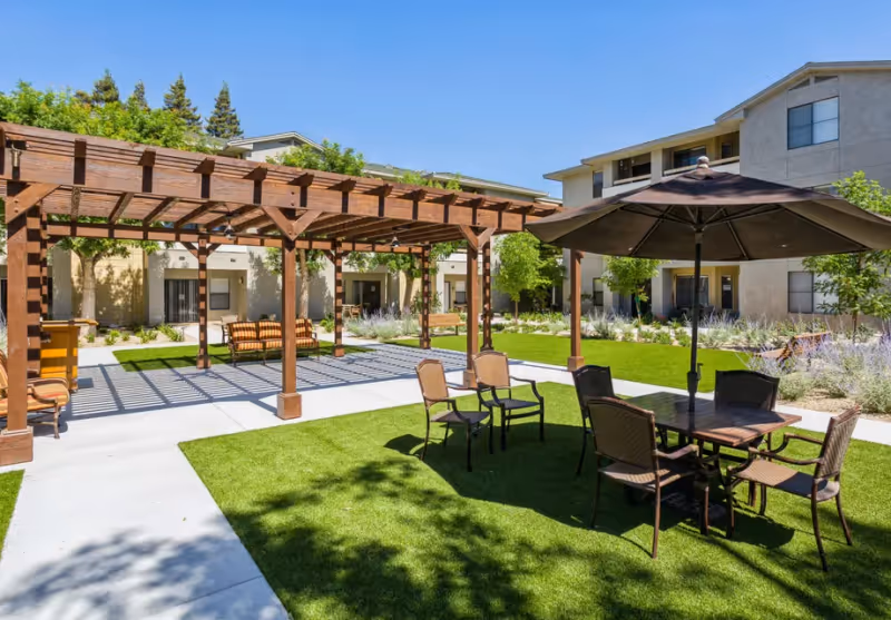 Outdoor seating area at a senior living facility with a wooden pergola, several chairs, a table with an umbrella, green grass, and surrounding buildings under a clear blue sky.
