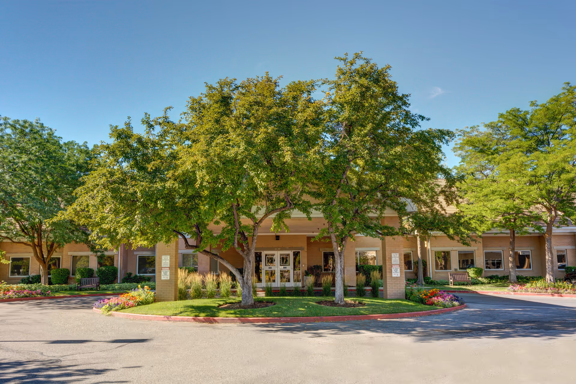 Front exterior view of The Auberge at Aspen Park facility with a circular driveway, green trees, and landscaped flower beds under a clear blue sky.
